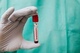 White gloved hand holding a clear vial labeled “coronavirus” with a red top. In the background is part of a blue disposable medical gown