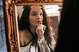 A woman puts on lipstick while looking in a mirror.