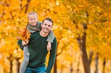 Father carries daughter through autumn trees. She holds yellow orange leaves.