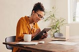 Photo of a woman in an orange shirt sitting at a table and taking notes on a tablet.