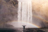 A man standing with his arm outsretched in front of a waterfall that’s creating a rainbow.