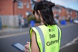 A person wearing a yellow high-visibility vest, printed with the words “Data Gatherer” and an Open Data Manchester logo, is standing on a residential street lined with red-brick houses, inputting data into a handheld electronic device.