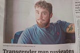 A photograph of a News&Observer newspaper shows a white trans man in North Carolina. He is blond, has a full beard, is wearing a blue t-shirt, and has a tattoo on his arm. The article in the newspaper is titled “Transgender man navigates anti-LGBTQ climate in NC”.