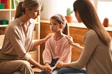 A warm and supportive scene in a classroom where a young girl, Sophie, is sitting with her mother and her teacher, who is gently explaining the importance of recognizing her emotions.