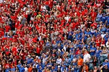 A stadium with fans wearing blue shirts in one section standing next to a section with fans all in red.