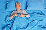 A photo of an older man asleep in a bed with blue sheets, pillowcases, and covers.