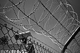 Black and white photo of a barbedwire fence and a security camera.