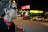 Night time in the Bronx, slightly blurred image of a man wearing a petite red nose and black Buddy Holly glasses in the foreground as he schleps past the blurred neon lights of the Riverdale Diner. He’s a medical clown on the way to a bat mitvah.