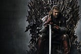 Eddard Stark sits on the Iron Throne. He gazes down. Both hands grip his sword, its point planted in the ground.