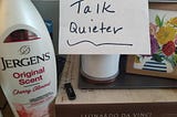 Picture of desk items including lotion, a candle, notecards and a handwritten sign that reads “talk quieter”.
