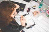 Young woman at her desk, looking at her diary while holding a pen. Photo taken from above.