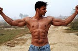 A dark, well-built, shirtless man poses on a dirt road. Wearing low-slung jeans, he shows off his biceps and six-pack.