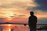 A silhouette of a person standing by a peaceful lake at sunrise, gazing at the sky. The sun casts a warm orange and pink glow over the water, creating a serene and reflective atmosphere.