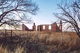 The crumbling brick remains of a building with two window outlines, two visible chimneys, and a missing roof framed between two small barren trees behind a barbed wire fence and in the middle of waist-high dead grass