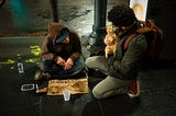 A man kneels on a sidewalk next to a homeless man, engaging him and an orange kitten in his arms. The homeless man seems amused by the man holding the kitten. Next to the homeless man is an open carry-out container with some partially eaten food, the spoon now resting in it. He also has a sign that says: “A lil helps a lot! Thank you! God Bless! Prayers please!”