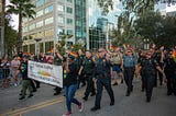 about a dozen uniformed police officers march in a parade, waiving rainbow flags, and holding a banner reading “central florida gay officers action league”