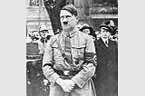 Hitler is standing in his uniform, hands folded.