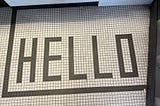 The image shows a tiled floor with the word “HELLO” spelled out in large, black, capital letters. The tiles are small and square, with a white background and black lines forming the letters and a border around the word. The word “HELLO” is centered within the border.