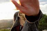 A person with long hair puts their hand out toward the viewer, blocking the view of their face.