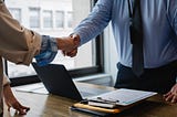 colleagues shaking hands in office over a desk with a laptop and some papers.