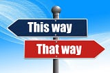 This is metal sign post with two directional arrows on it. On top is a blue one with the words, “This way” (pointing to the left), and below it is a red one with the words, “That way” (pointing toward the right). The background is sky blue with swishes coming down diagonally from top left to bottom right.