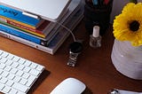 Organised desktop clutter in a home office: MacBook, magic mouse and keyboard, design books, flowers, pen pot full of markers, cuticle oil, and a smart watch on charge