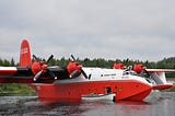 A very large red and white four engined amphibious aircraft floating in a lake with a rowboat beside it.