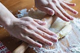 Two hands rolling out dough with a wooden rolling pin on a wooden table covered in flour.