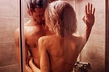 A sexy young woman with platinum blonde hair and a well-built, dark haired man with a beard embrace in a shower.