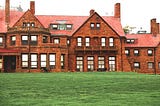 Large estate building with 4 chimneys and 20 windows visible on the two stories. Constructed of brick with two balconies and a large green lawn.