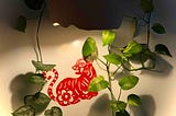 A red paper‑cut cat with floral patterns hangs on a light wall, surrounded by trailing green pothos vines that frame the artwork with soft, natural curves.