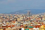 A colorful city spread out with low rise buildings — in the distance a brown cathedral under construction towers over the city. A hazy mountain is visible in the background.