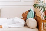 A woman in a white top with a light turquoise reclines on a white couch with her eyes closed.