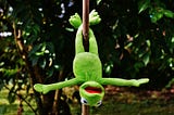 Kermit the frog hangs upside down on a pole by his feet in the garden.