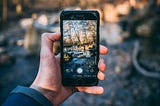 A hand holding an iPhone that is in photo-taking mode with an image of the sun-lit river and forest behind the iPhone.