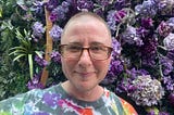 The author is standing in front of a wall of purple flowers and leaves. His hair is cut very short, and he is wearing brown glasses. He is smiling slightly, but it looks a little forced. He is wearing a colorful tie dye shirt.