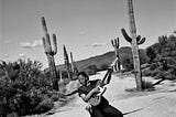 Beverly “Guitar” Watkins holding her guitar “Red Mama” in the middle of the dessert surrounded by cactuses and mountains.