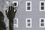 A black and white photo showing a person’s hand resting against a window with rain droplets on it.