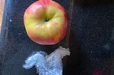 A red and yellow apple sits on a counter. A wad of plastic wrap sits next to it.