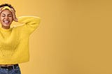 Carefree, happy, attractive girl in a yellow sweater and headband, touching head and smiling broadly. Yellow background.