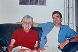 Man in blue shirt and older woman sitting on a couch smiling