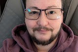 selfie: Grayson in a dark hoodie, wearing glasses, short hair parted, with a beard