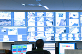 A transit operator works diligently with their console and a video wall in a dimly lit room.