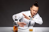 A man in a dress shirt and time sitting at a dinner table shelters himself from the glass and bottle of scotch in front of him that is shackled to his wrist