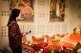 A visitor, side profile, wearing headphones and holding a smartphone, is focused on an audio guide. They stand before a vibrant exhibit of traditional red and gold musical instruments, with a large mural in the soft-focused background.