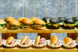There are two wooden shelves of burgers and hotdogs. On the top shelf, on the left are chicken burgers consisting of a sesame seed bun, split in two, each containing a breaded, fried chicken fillet, a slice of cheese and lettuce. On the right of the top shelf are black charcoal buns, each containing a beef patty topped with a slice of melted cheese and some lettuce. The bottom shelf contains split hot dog buns, filled with hotdogs and pink coleslaw, each in a little paper tray.