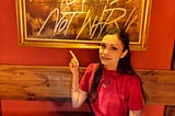 Woman in red shirt sitting at wooden table, smiling and pointing up at framed golden artwork behind her that reads ‘Make Cocktails Not War.’