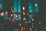 A picture of many hanging lightbulbs burning brightly against the backdrop of a dark room partially illuminated by light coming through blue curtains.