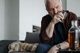 Image shows older man looking frustrated and upset while drinking whiskey from a tumbler while bottle of whiskey sits on a table in the foreground.