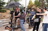 Seven students and one instructor cluster around a cart-mounted camera on a location shoot for Eight & Sand/photo by Avery Jackson